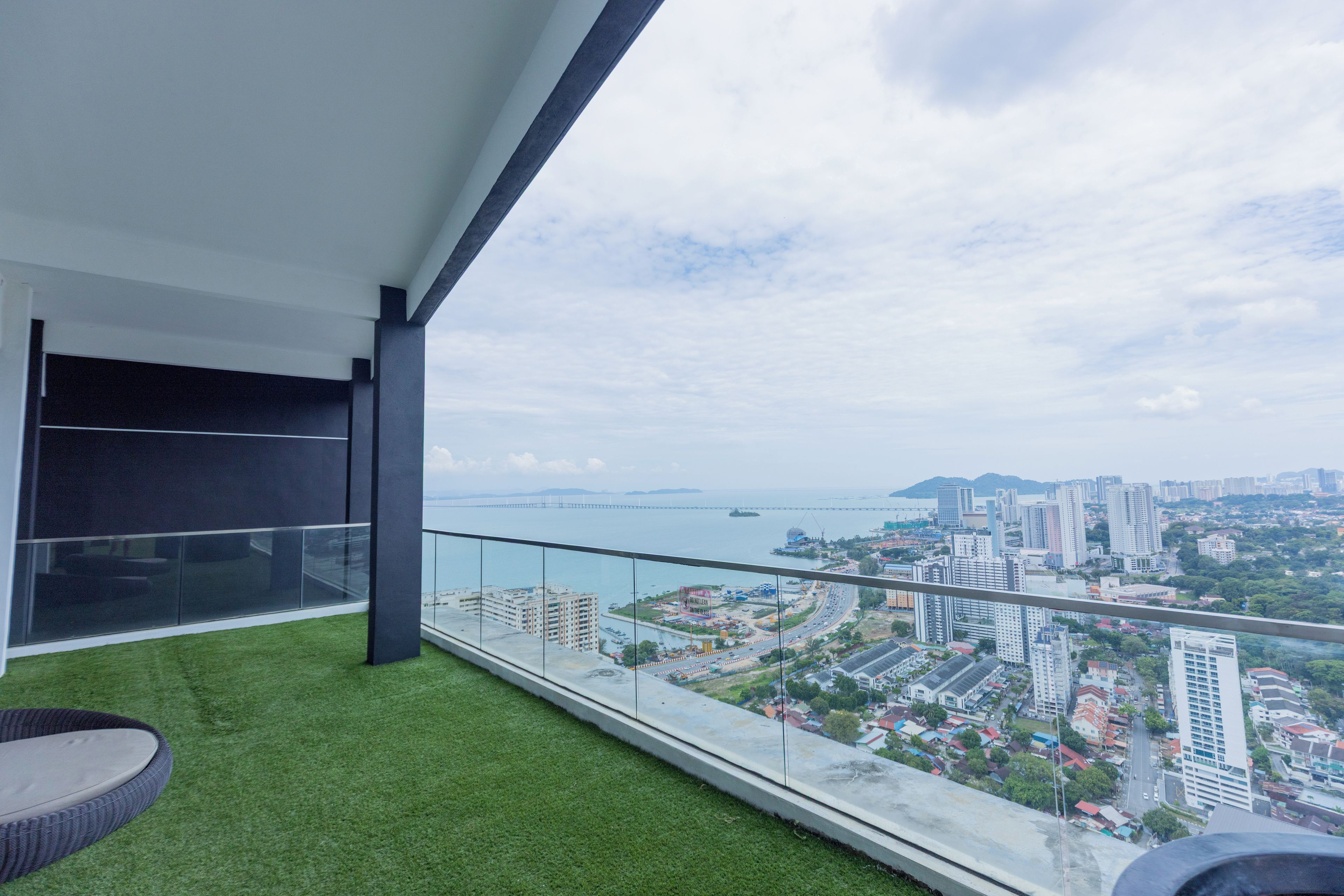 A balcony with artificial grass, a glass railing, and a view of a cityscape and water body.