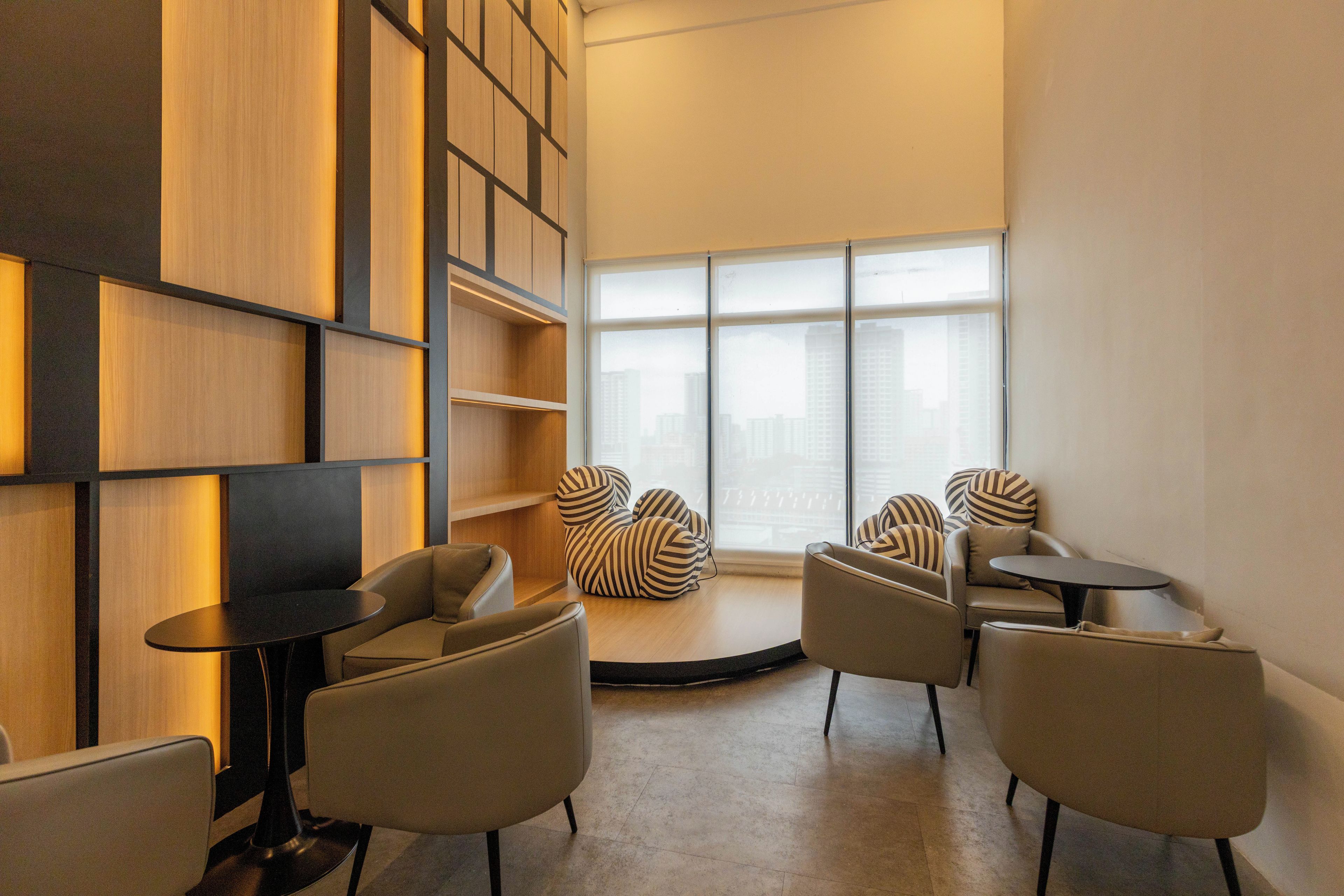 A modern lounge area with a circular seating arrangement, a large window with a city view, and built-in wooden shelving.