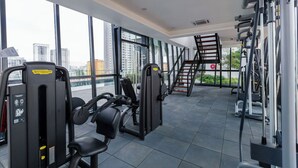 Une salle de sport équipée de divers appareils d’exercice, dotée de grandes fenêtres et offrant une vue sur le paysage urbain.