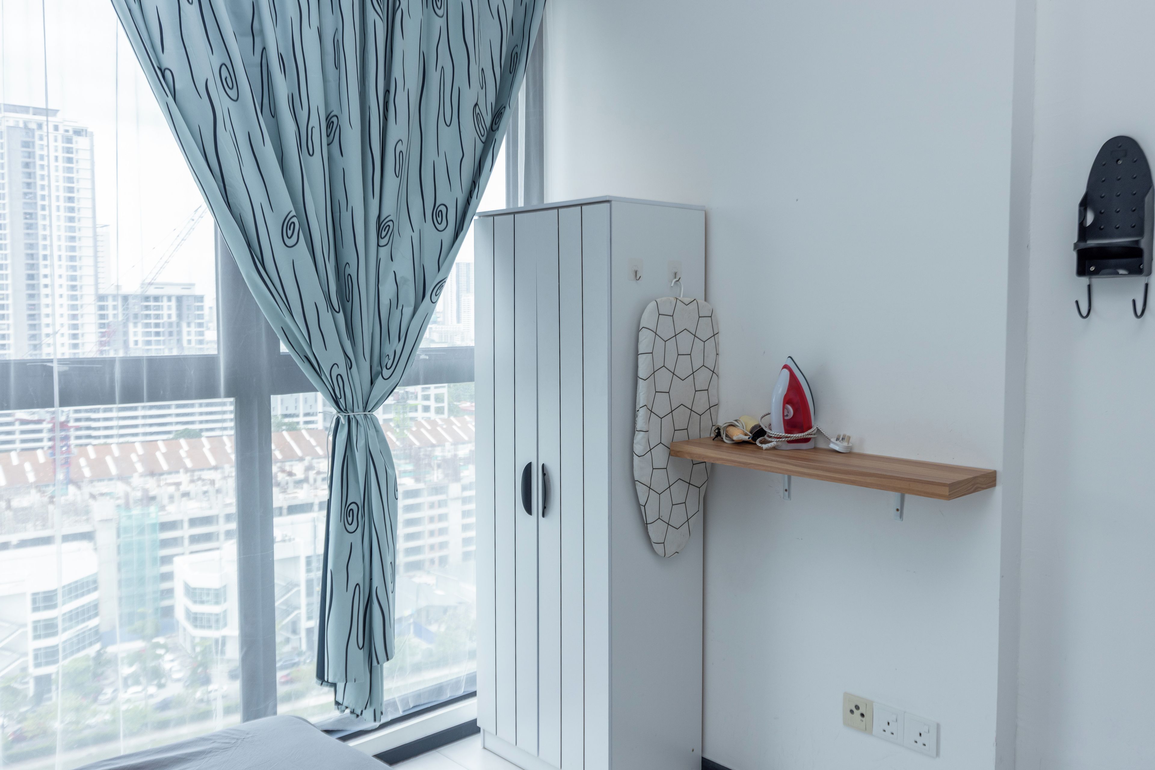 A room with a wardrobe, a shelf with an ironing board and iron, and a window with a curtain.
