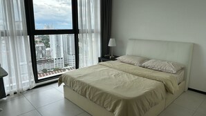 Une chambre avec un grand lit, une table de chevet avec une lampe, et une vue sur de hauts immeubles à travers des rideaux légers.
