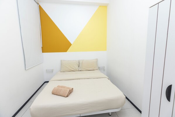 Ein Einzelbett mit beiger Matratze und Kissen, ein aufgerolltes Handtuch und eine geometrische Wanddekoration.