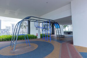 An outdoor playground with a climbing structure and a slide, surrounded by a landscaped area with trees and shrubs.