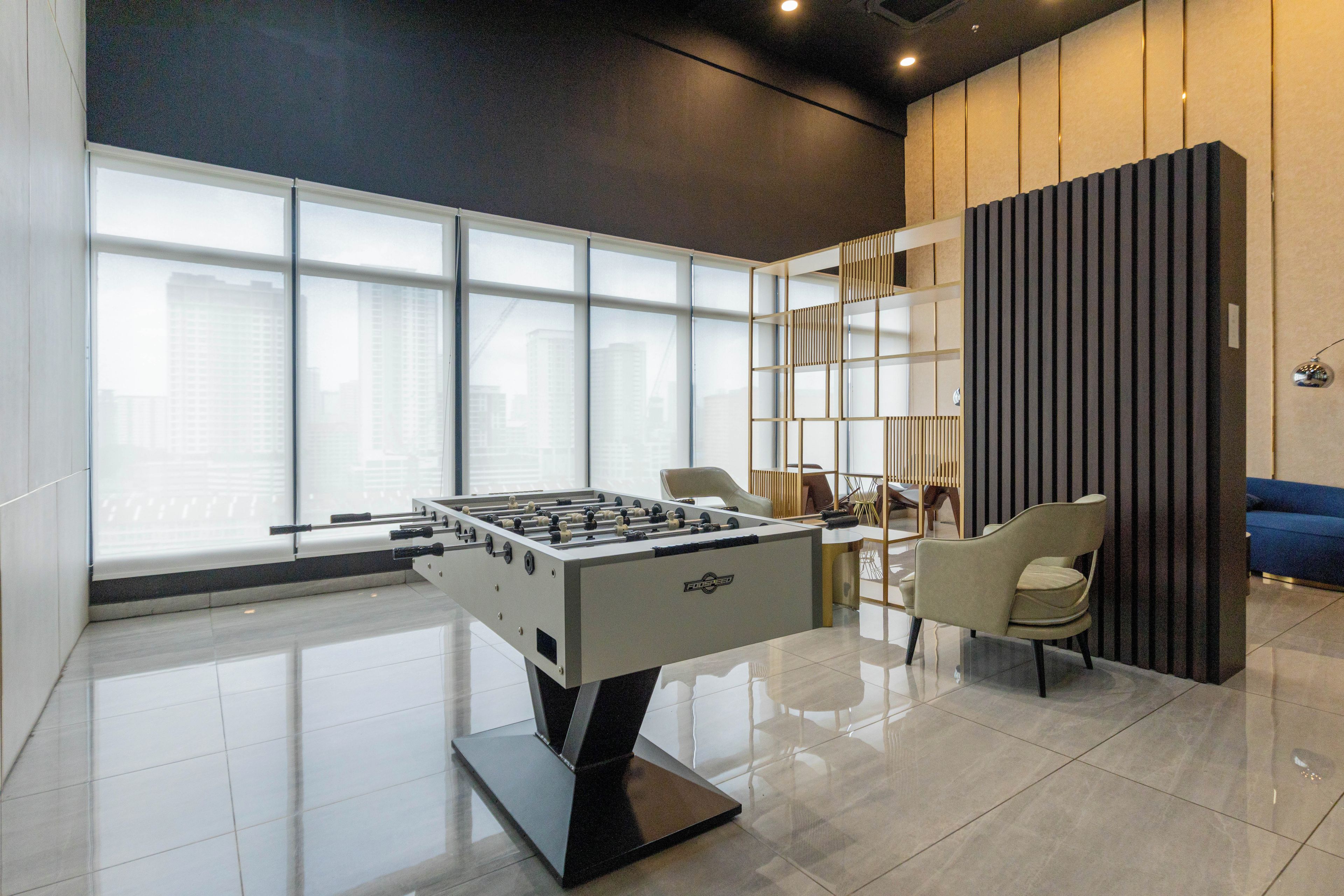 A modern indoor space with a foosball table, a seating area with chairs, and a large window providing a view of buildings outside.