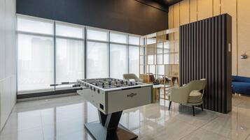 A modern indoor space with a foosball table, a seating area with chairs, and a large window providing a view of buildings outside.