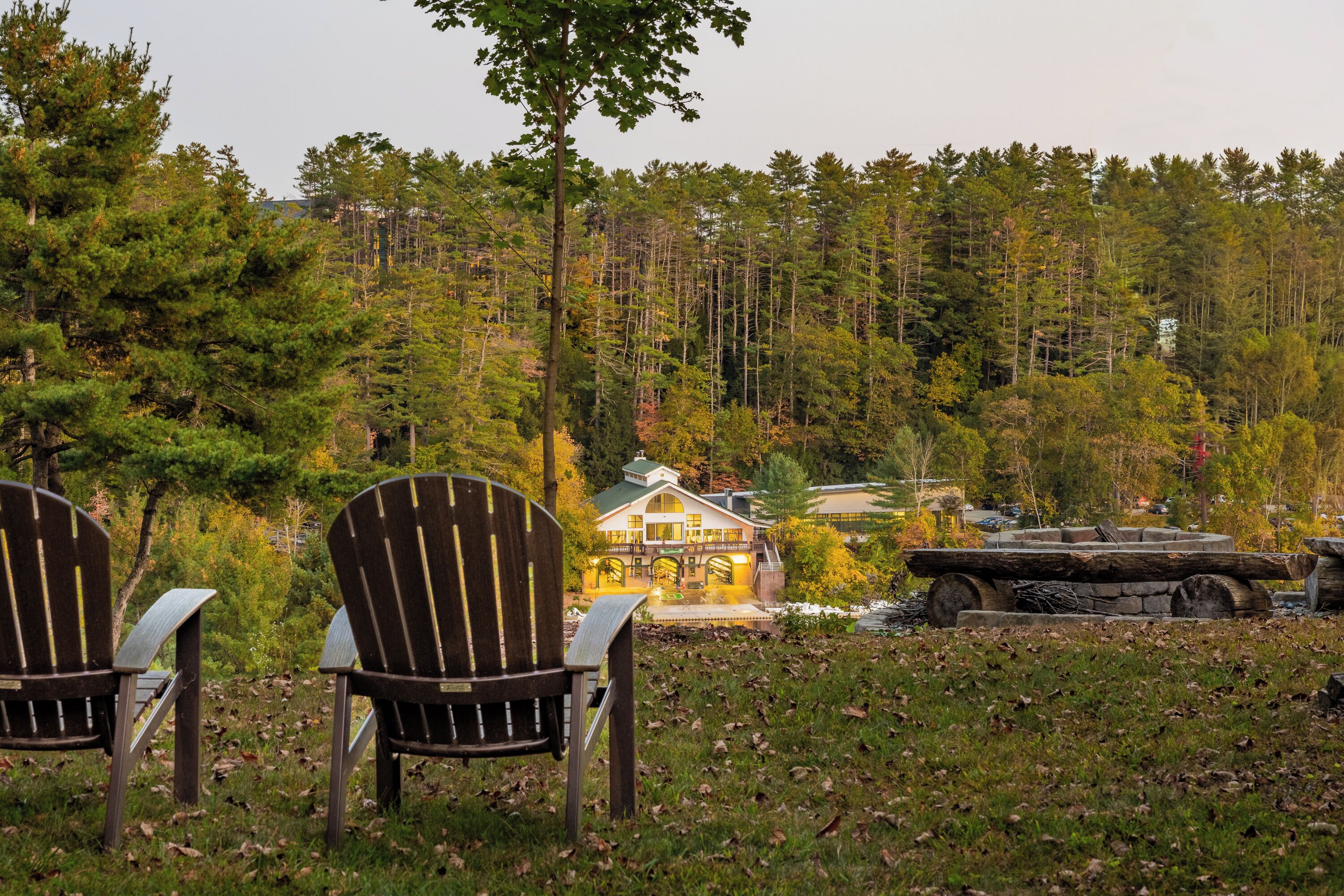 Two Adirondack chairs facing a lodge with a stone fireplace and a pond.