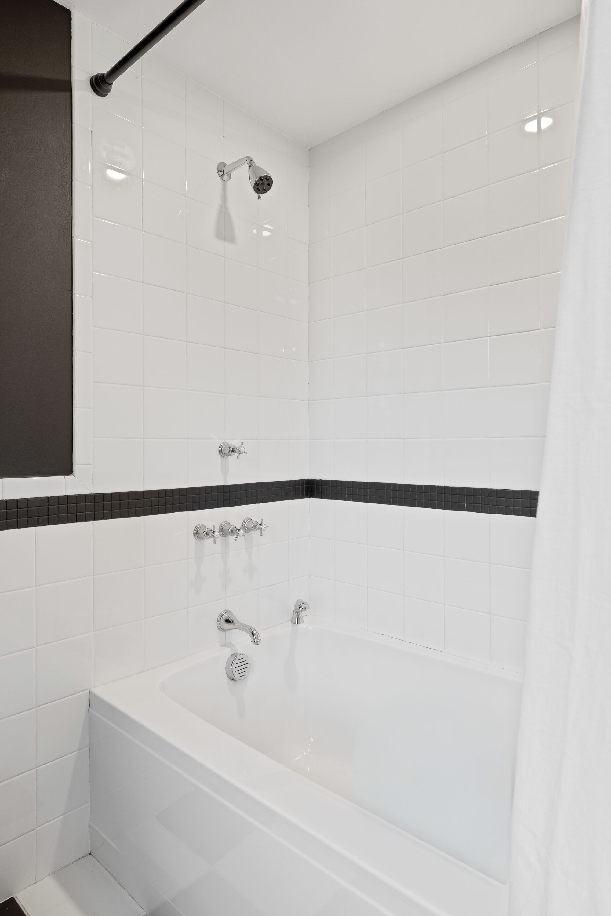 Ein modernes Badezimmer mit weißen Fliesen, einem schwarzen Akzentstreifen aus Fliesen, einer Badewanne und einem Duschkopf.