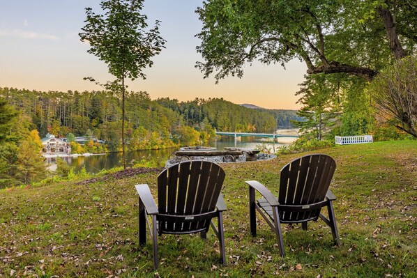 Två Adirondack-stolar placerade framför en sjö, med en bro och ett hus i bakgrunden.