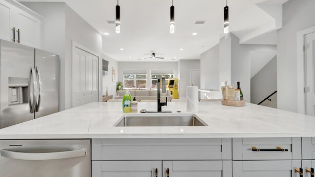 Eine moderne Küche mit weißen Schränken, einem Edelstahlkühlschrank, einer großen Kochinsel mit Spüle, Pendelleuchten und einem Deckenventilator.