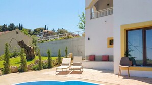 A swimming pool with lounge chairs, a white building with balconies, and a view of the surrounding landscape.