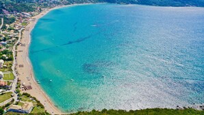 An aerial view of a coastal town with a beach, clear turquoise waters, and lush green hills in the background.