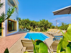 A swimming pool with lounge chairs, a white building with a window, and greenery in the background.