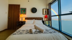 A hotel room with a bed, two bedside tables, a door, and a large window with a view of the sea and a rainbow. - 康宿·海映居 | KanStay Oceanview Residence 4005 | 2BR 2BA · Manila Bay View (Manila)