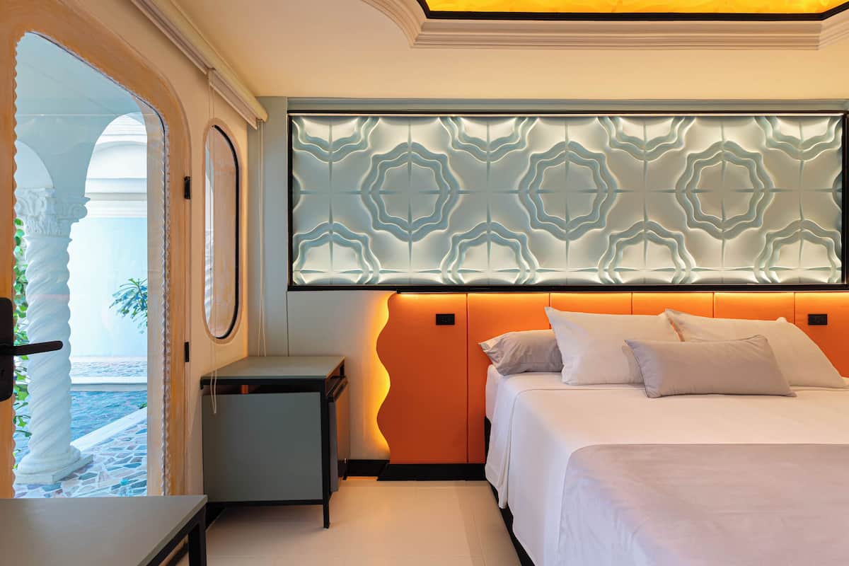 Une chambre d’hôtel avec un lit, une table de chevet, une tête de lit décorative et une vue sur la mer, visible à travers une porte en arc de cercle.