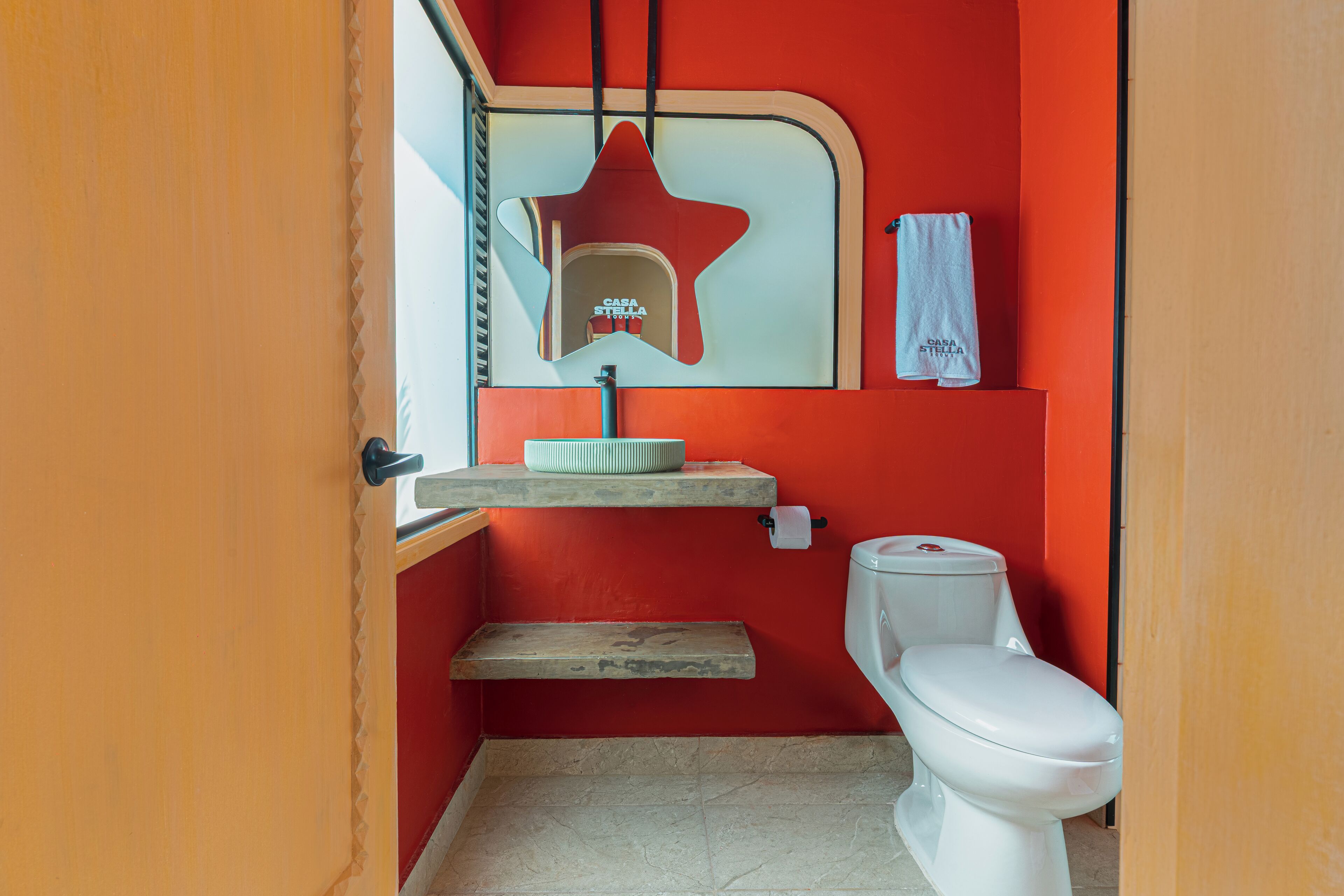 Une petite salle de bain avec un mur rouge, des toilettes blanches, un petit lavabo avec un miroir au-dessus, et une serviette accrochée au mur.