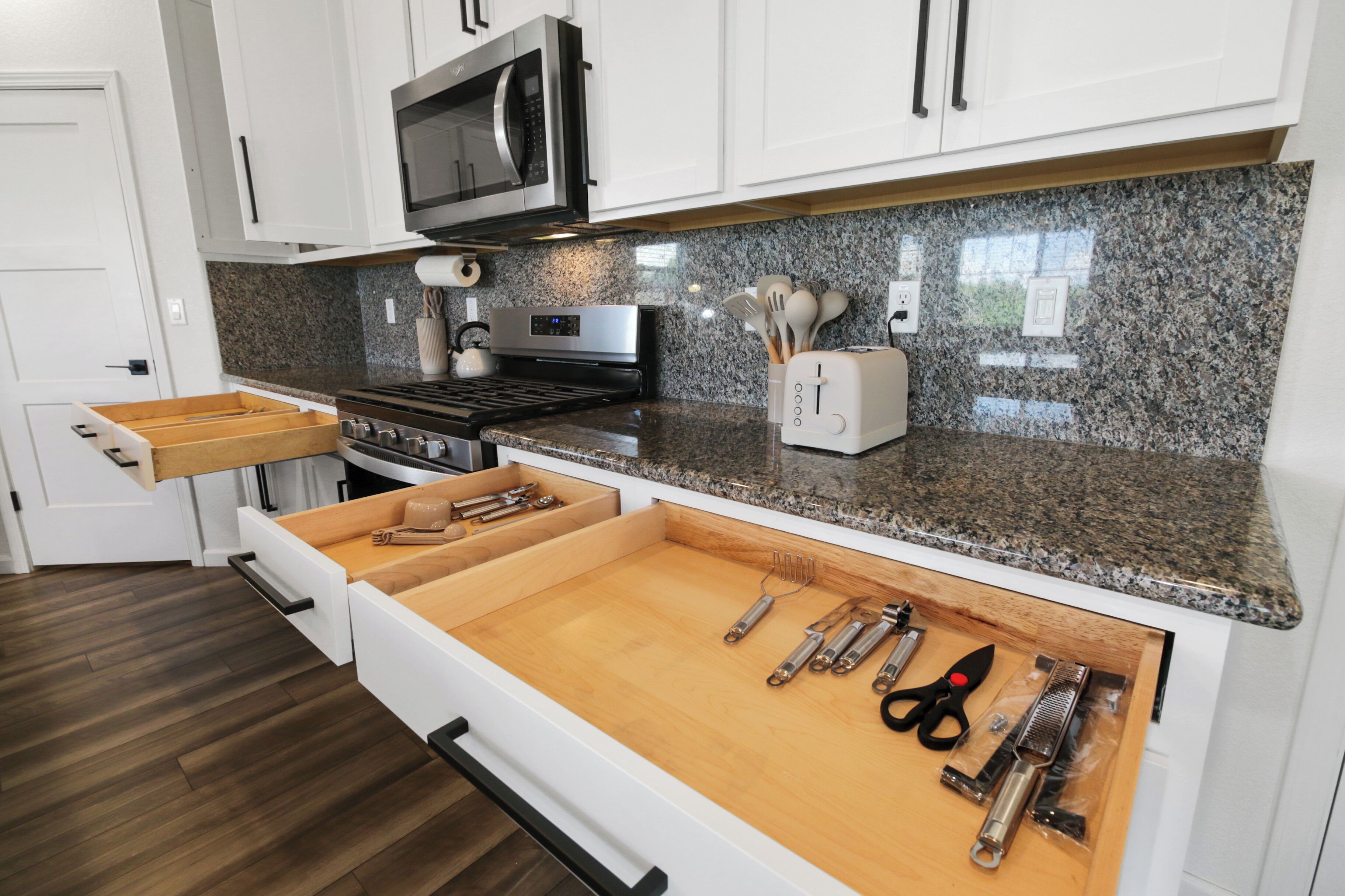 A kitchen with white cabinets, a granite countertop, a built-in microwave, and a drawer organizer with utensils.