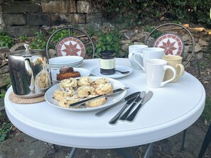 A round outdoor table with a white tablecloth, a metal kettle, a plate of scones, a cup of coffee, a mug, a jar of jam, and a slice of cake.