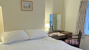 Una cama bien hecha con ropa de cama blanca, una mesita de noche con lámpara, una silla con cojín escocés y un cuadro en la pared.