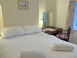 Un lit bien fait, avec une literie blanche, une table de chevet avec une lampe, une chaise dotée d’un coussin à carreaux et un tableau encadré accroché au mur.
