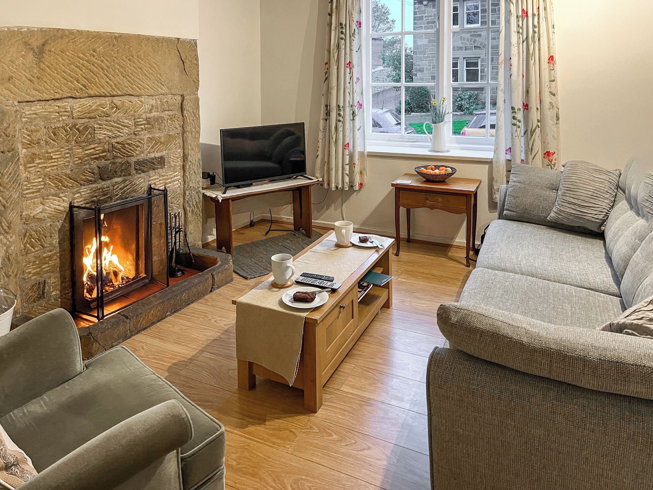 A cozy living room with a stone fireplace, a television on a wooden stand, a sofa, a coffee table, and a window with curtains.