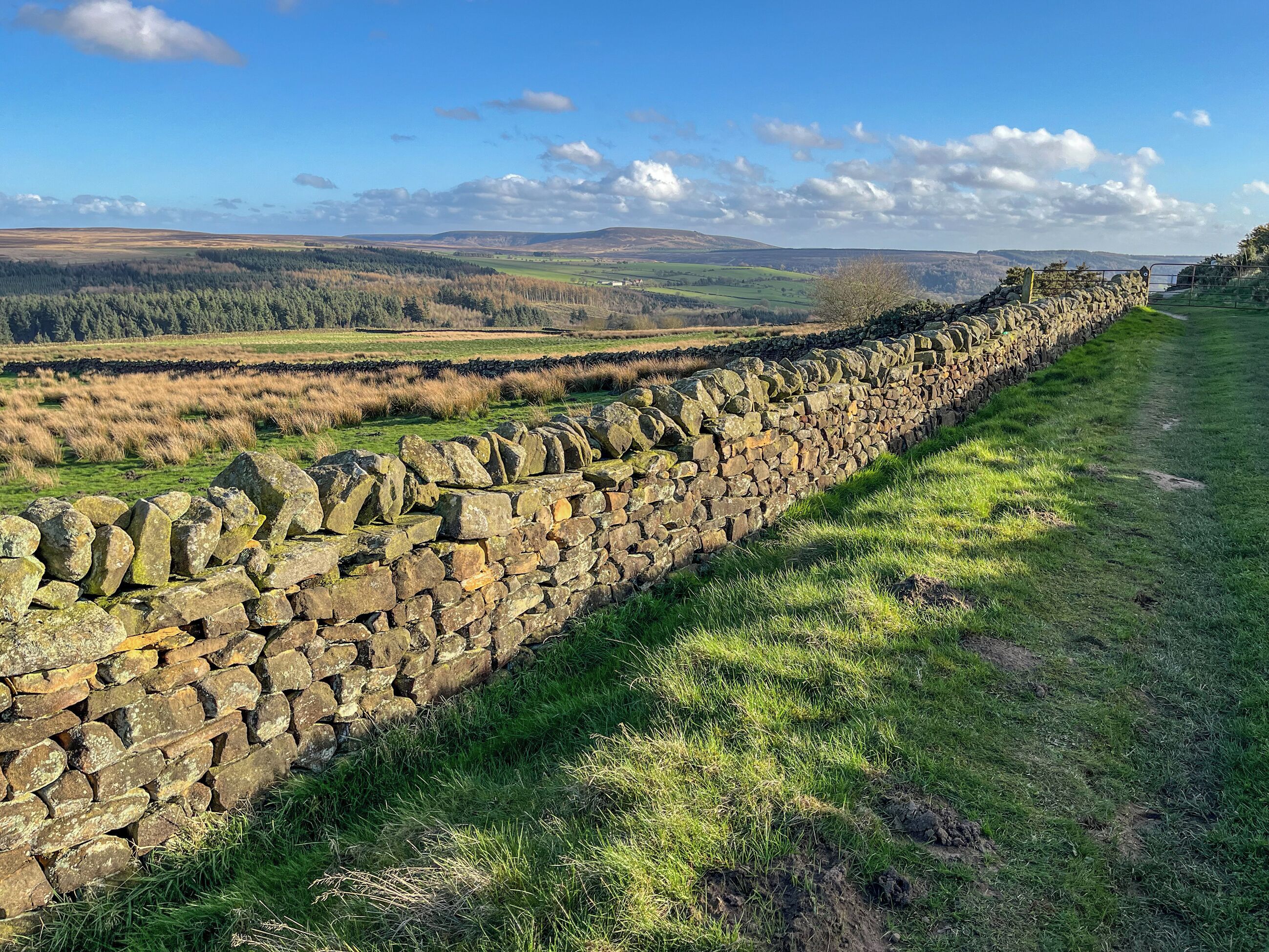 A stone wall with a grassy path beside it, leading to a scenic view of rolling hills and a clear sky.