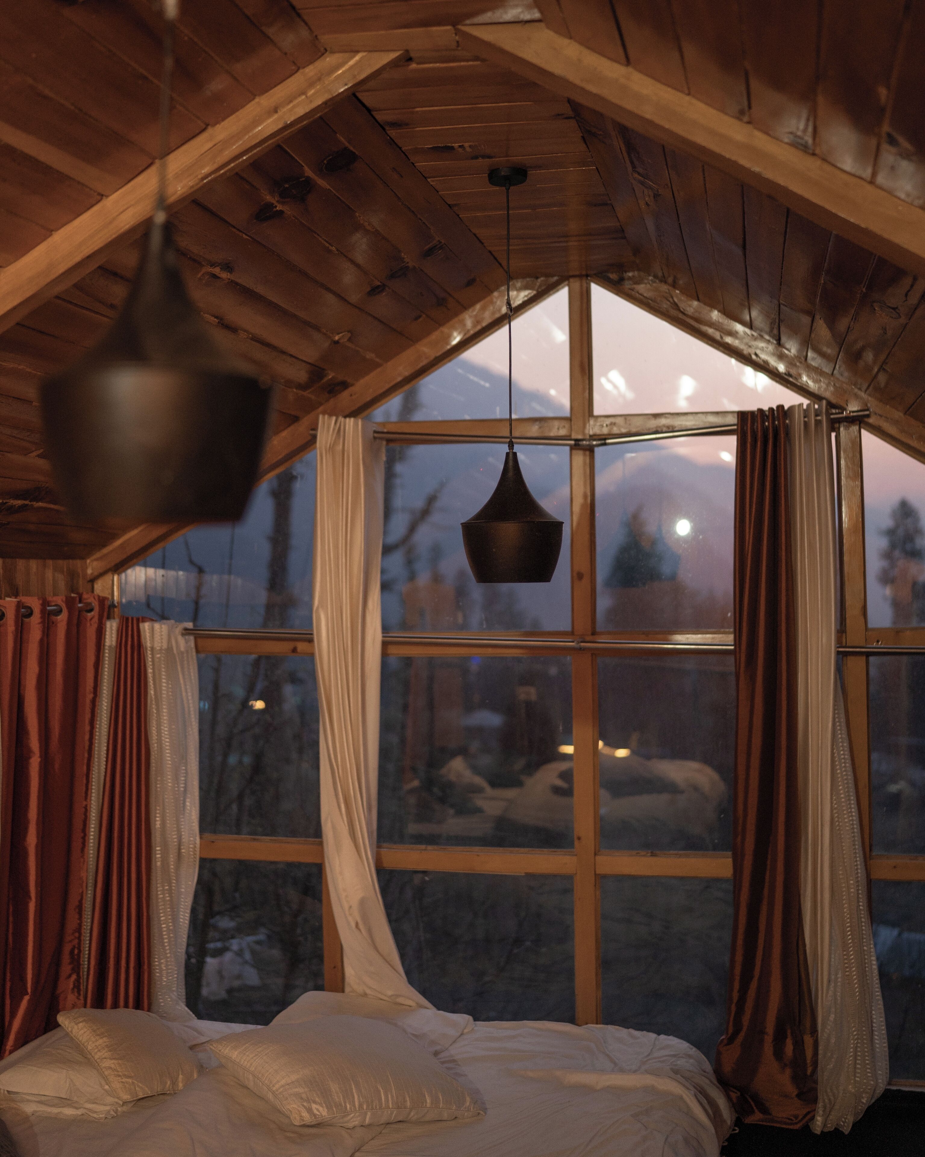 A room with a bed, wooden ceiling, large window, and curtains.