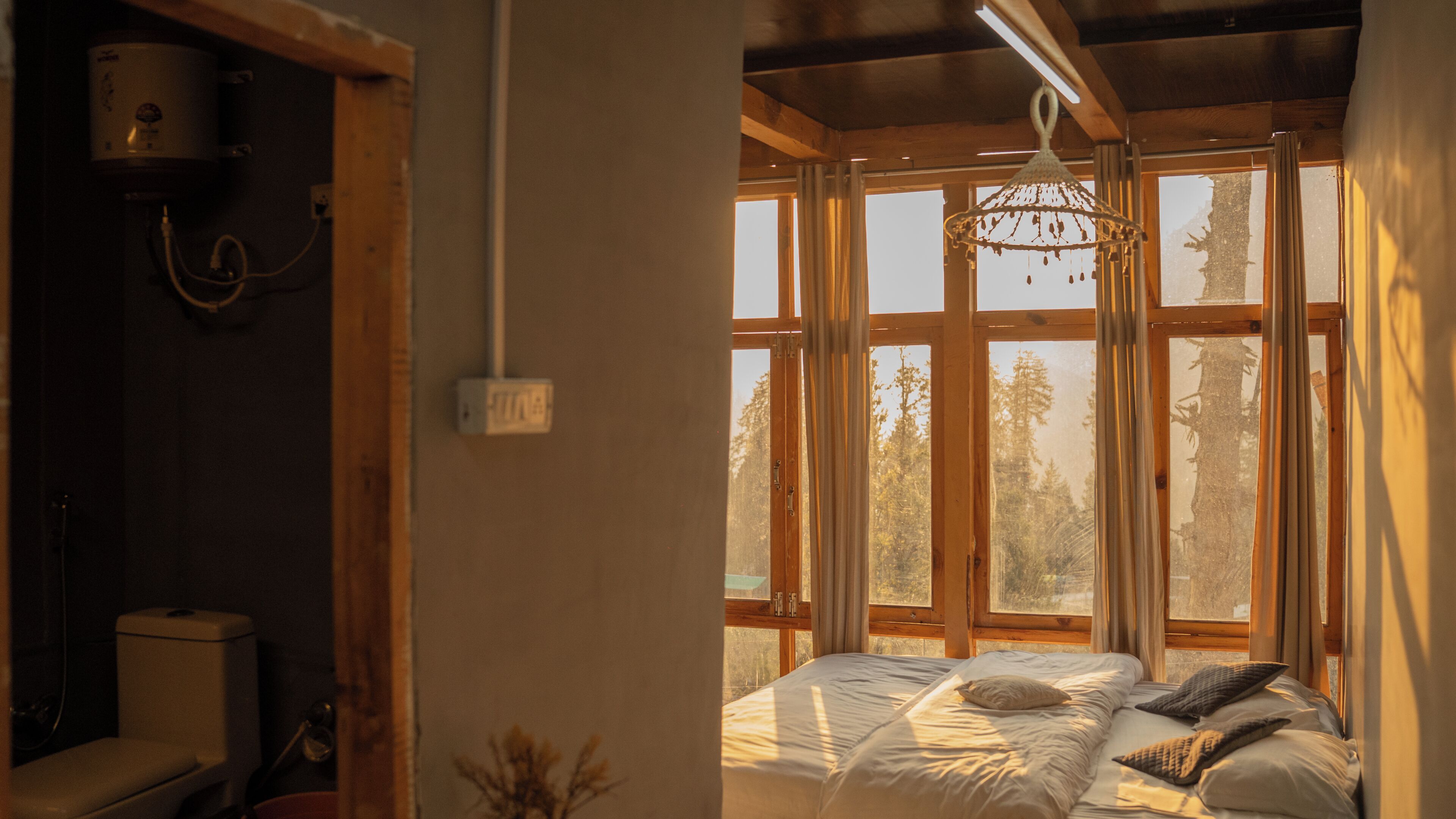 A bedroom with a bed, a window with curtains, a hanging light fixture, and a visible electrical outlet.