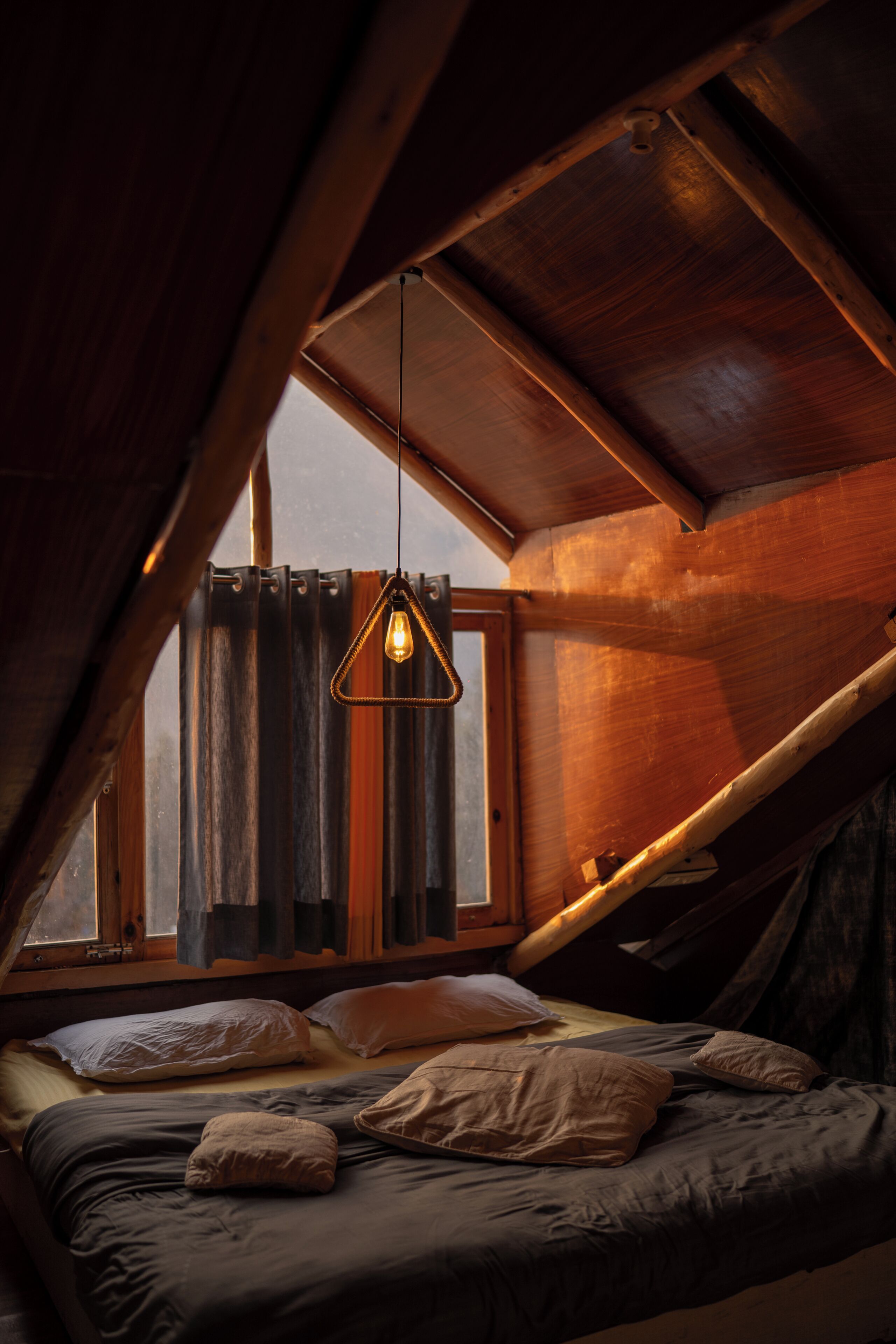 Un dormitorio con techo inclinado, lámpara colgante, cama con almohadas y ventana con cortinas.