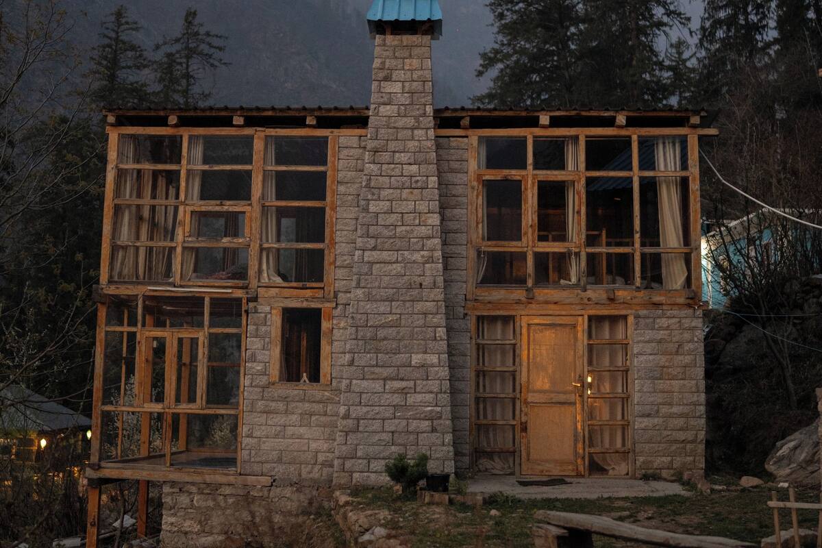 Un edificio de piedra con ventanas amplias y una chimenea, rodeado de árboles y montañas.