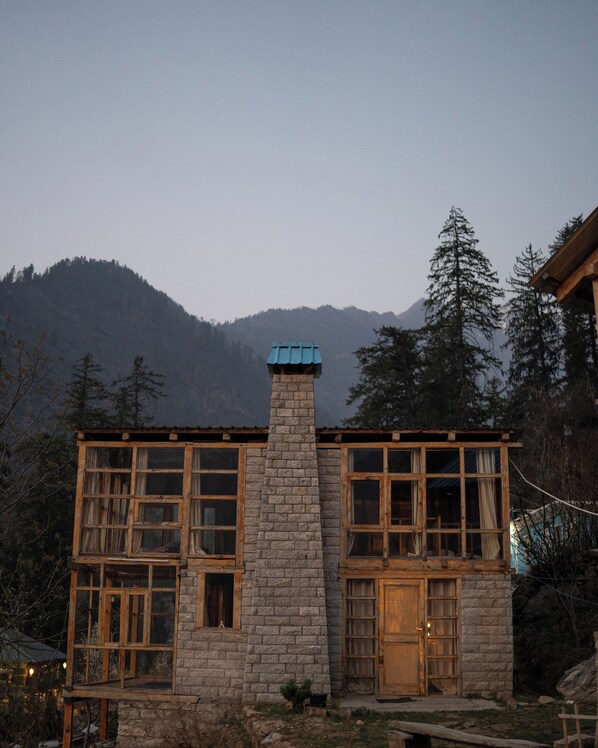 A stone building with large windows and a chimney, surrounded by trees and mountains.