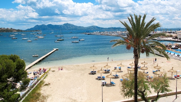 A beach with clear blue water, boats, and a mountain in the background.