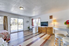 Une pièce spacieuse dotée d’une grande porte-fenêtre coulissante donnant sur un balcon, d’un bureau blanc avec une chaise, d’un téléviseur fixé au mur et d’un coin bar avec des tabourets.