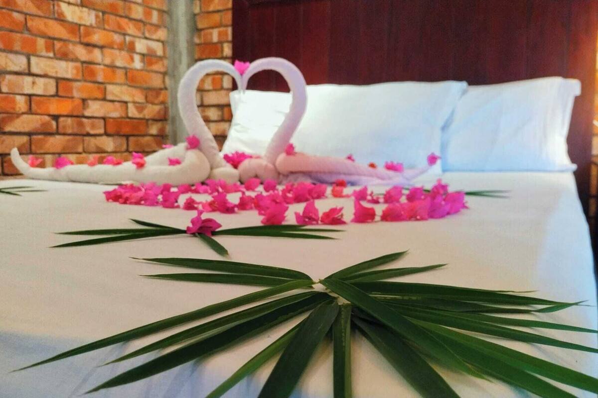 Una cama cuidadosamente tendida con una decoración en forma de corazón hecha con flores y cisnes, una hoja de palma y un muro de ladrillo al fondo.