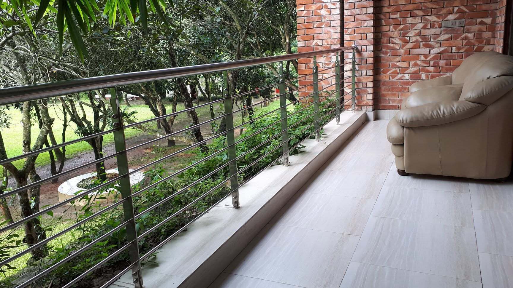 A balcony with a metal railing, a beige sofa, and a view of a garden with trees and a circular structure.
