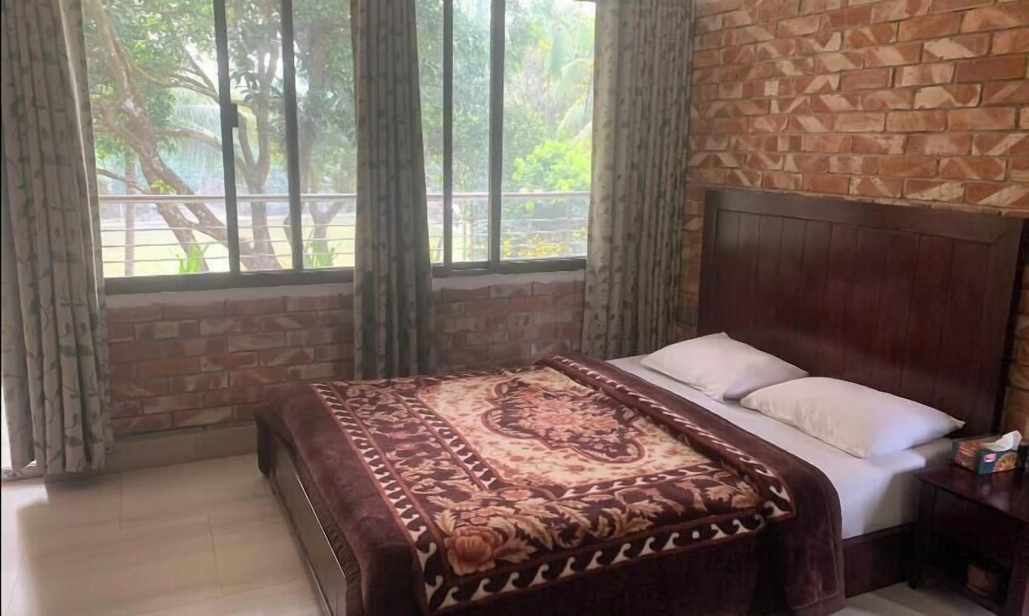 Un dormitorio con pared de ladrillo, una ventana grande con cortinas, una cama con colcha estampada y una mesita de noche con un cajón.