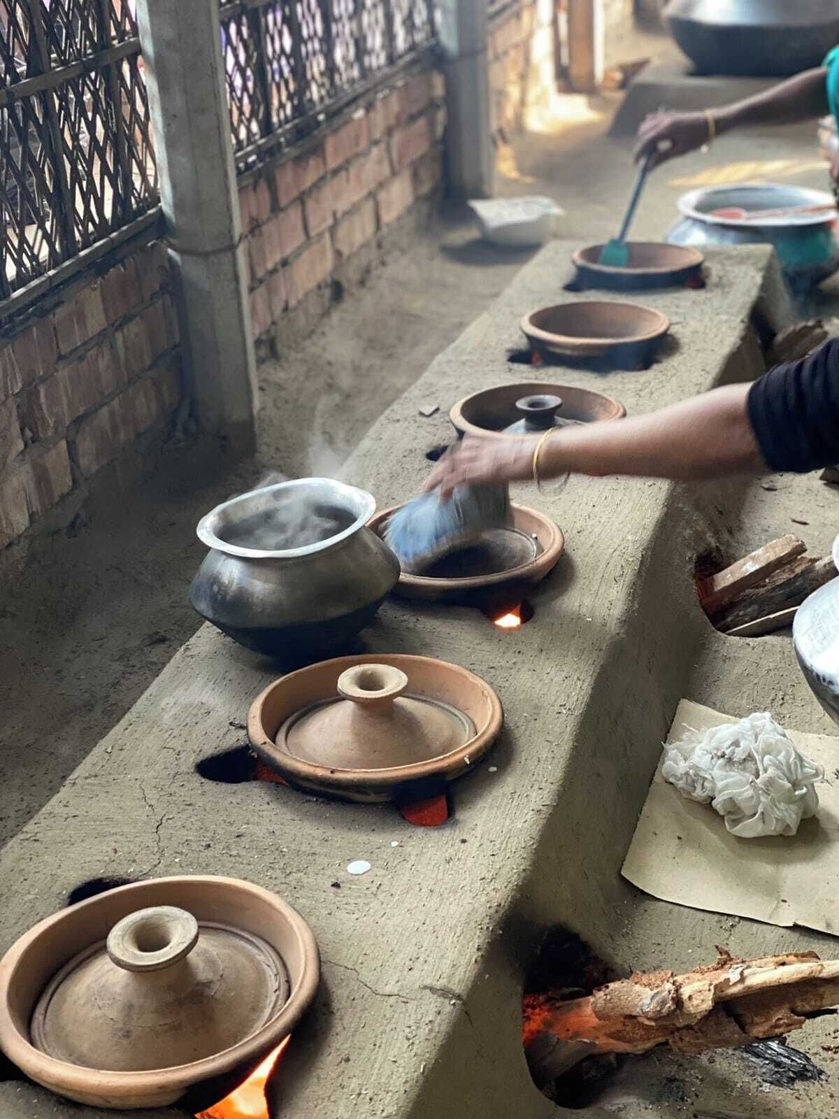 A row of clay pots on a concrete ledge, some with lids and others open, with a person tending to the fire below.