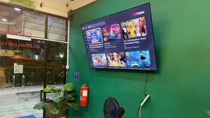 Ein digitaler Bildschirm zeigt Musikvideos in einem Eingangsbereich mit grüner Wand, Feuerlöscher, Ventilator und Willkommensteppich.