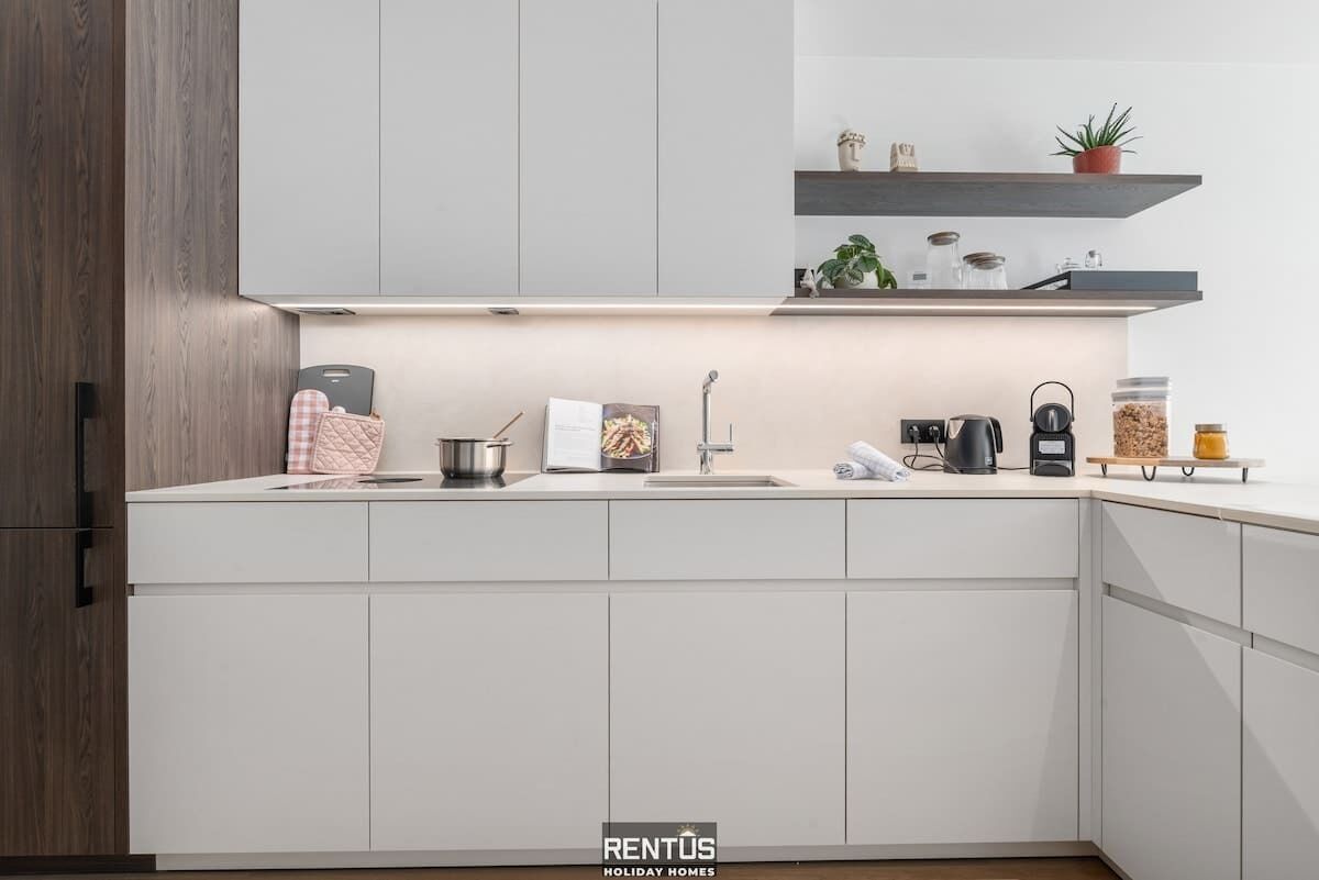 A modern kitchen with white cabinetry, a built-in oven, a sink, and a coffee maker. There are shelves above the counter holding decorative items and plants.
