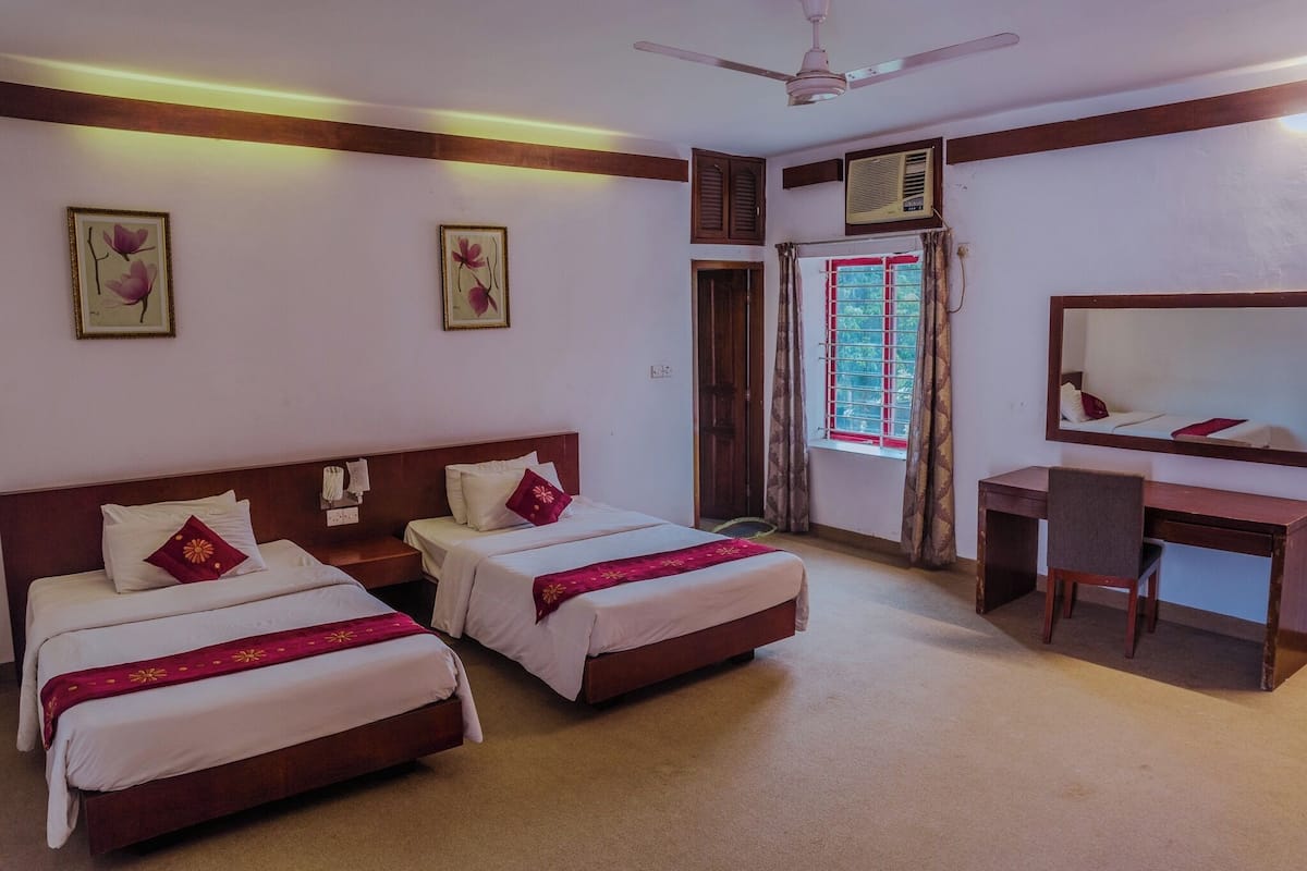 A hotel room with two single beds, a desk, a chair, a ceiling fan, an air conditioning unit, and a window with curtains.