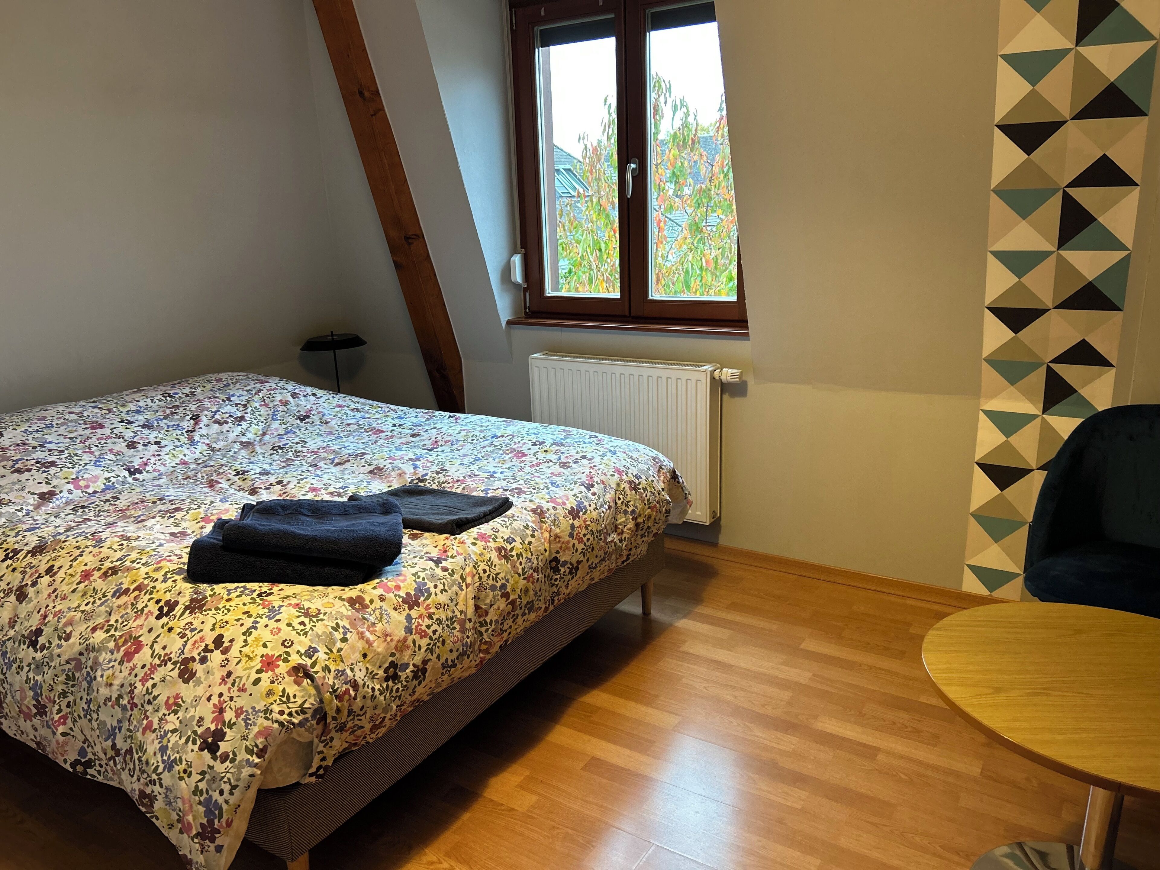 Une chambre à coucher avec un lit, un radiateur, une fenêtre donnant sur des arbres, un plancher en bois et une chaise.