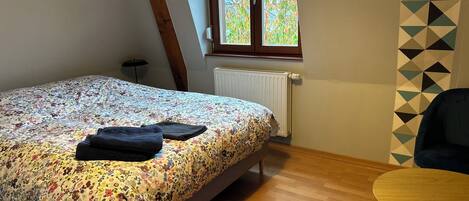 Une chambre à coucher avec un lit, un radiateur, une fenêtre donnant sur des arbres, un plancher en bois et une chaise.
