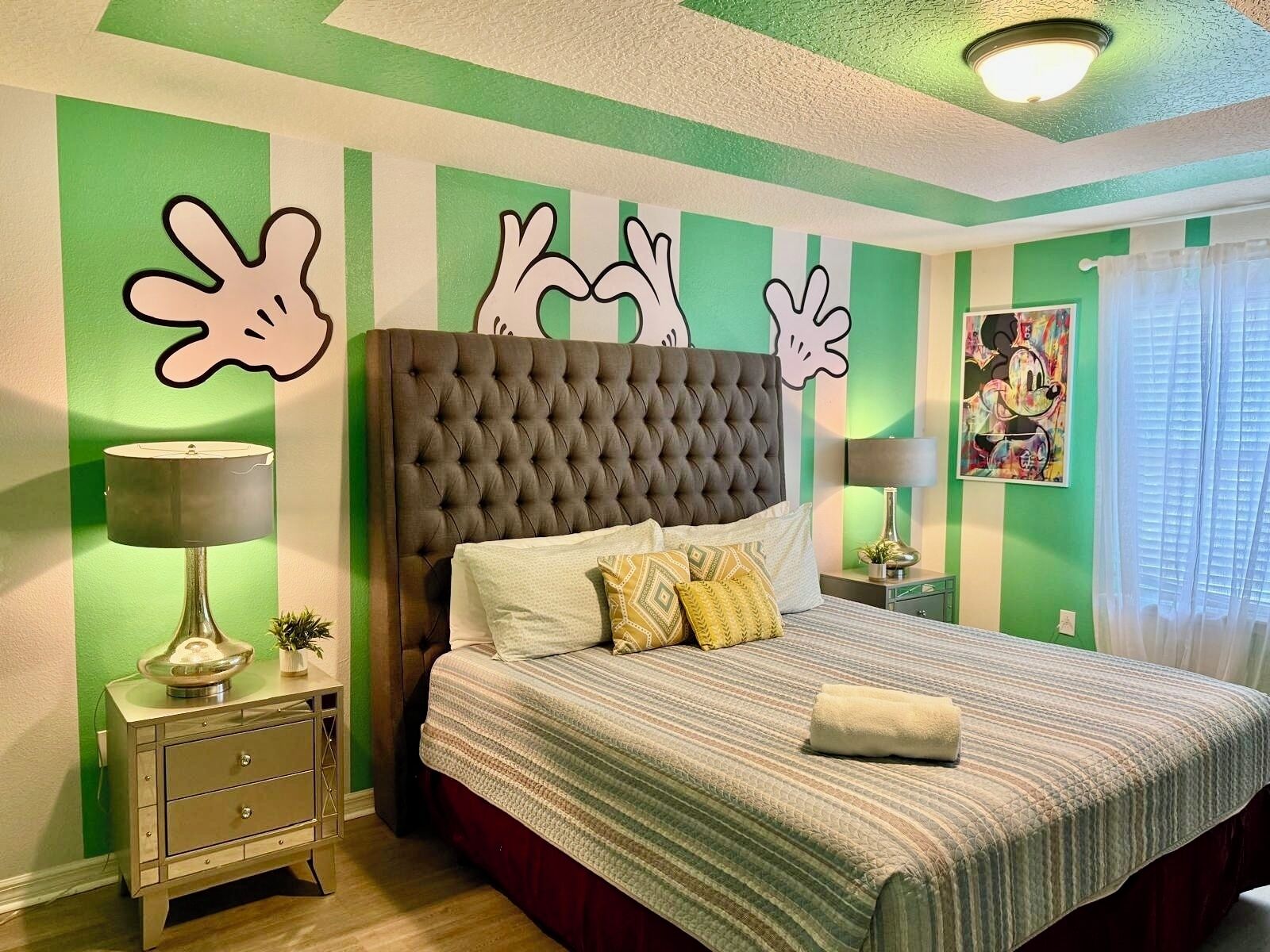 A bedroom with a tufted headboard, a bed with striped bedding, two bedside tables with lamps, and a green-striped wall with abstract art.