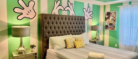 A bedroom with a tufted headboard, a bed with striped bedding, two bedside tables with lamps, and a green-striped wall with abstract art.