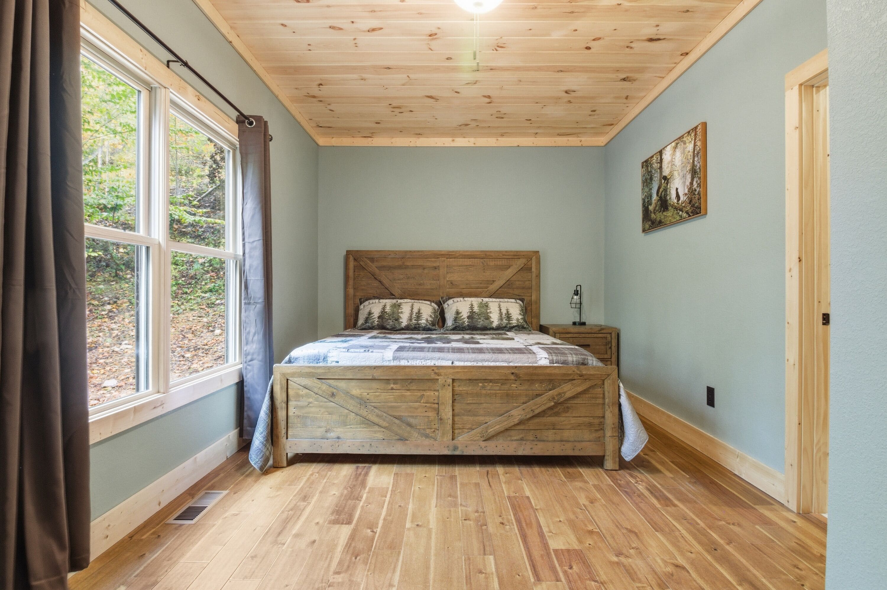 A bedroom with a wooden bed, a large window with curtains, a painting on the wall, and a wooden ceiling.