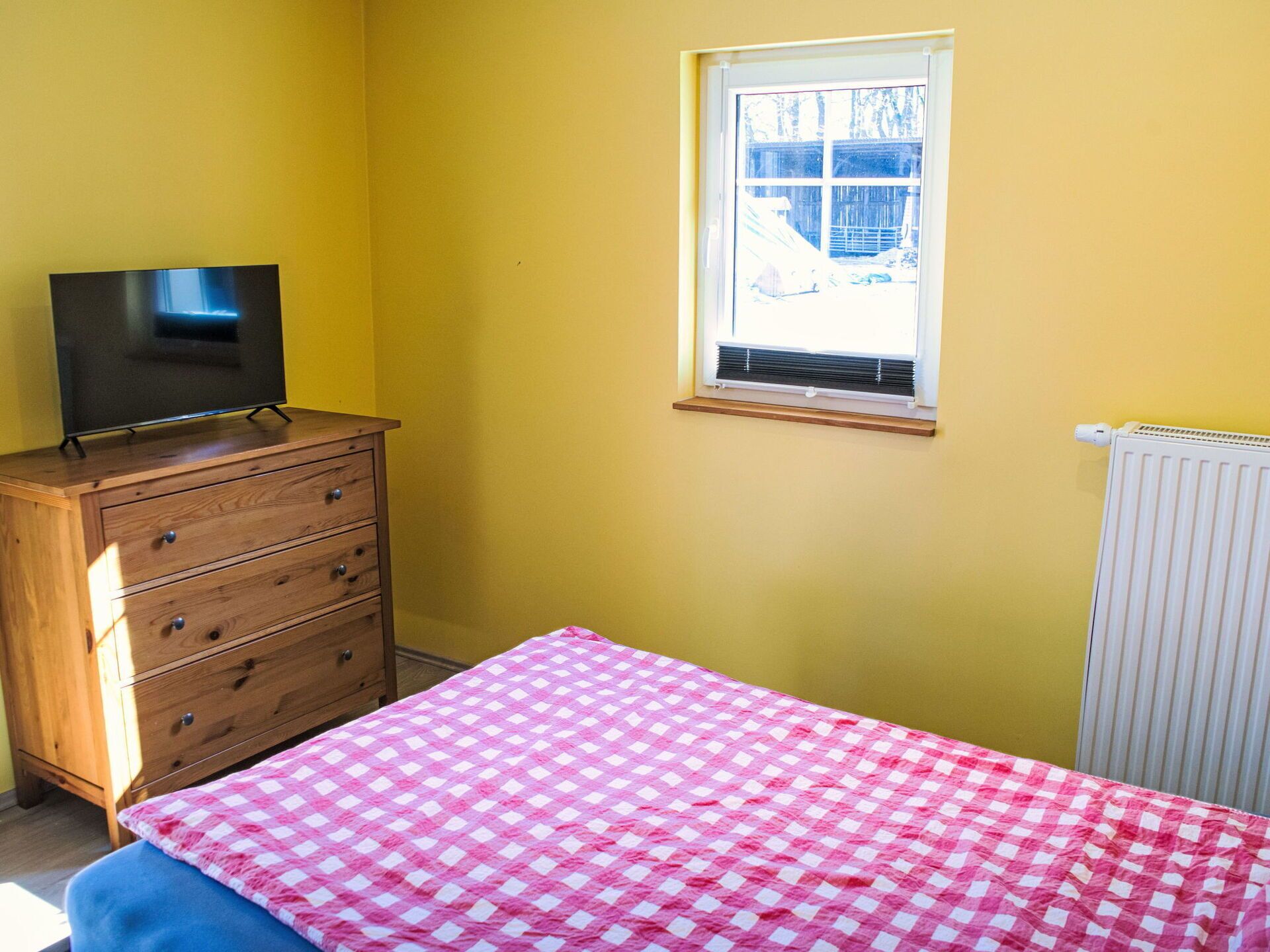 Una habitación con cama, cómoda, televisor encima y ventana con vistas, además de un radiador.