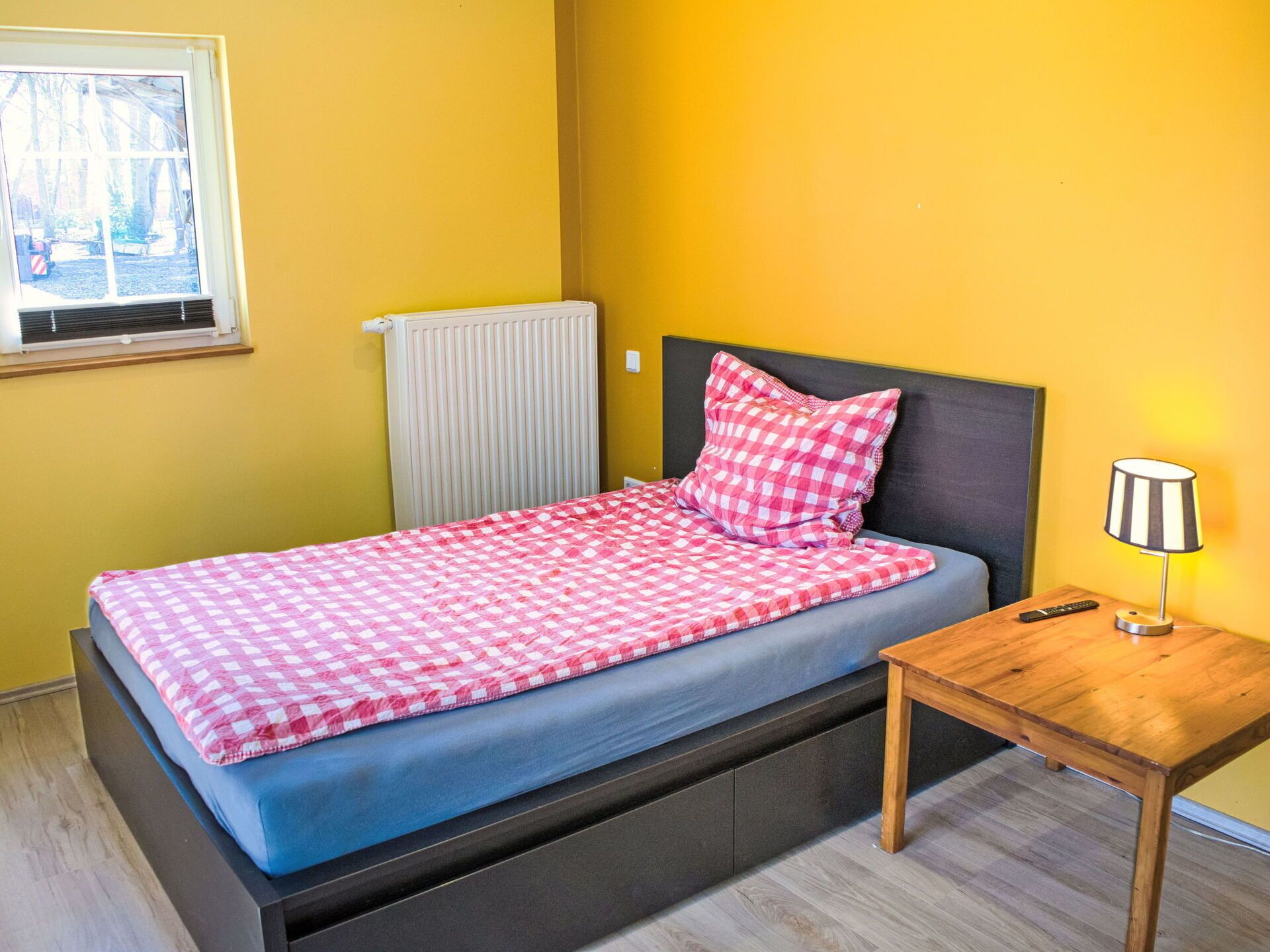 A single bed with a checkered bedspread and a pillow, a bedside table with a lamp, a radiator, and a window with blinds.