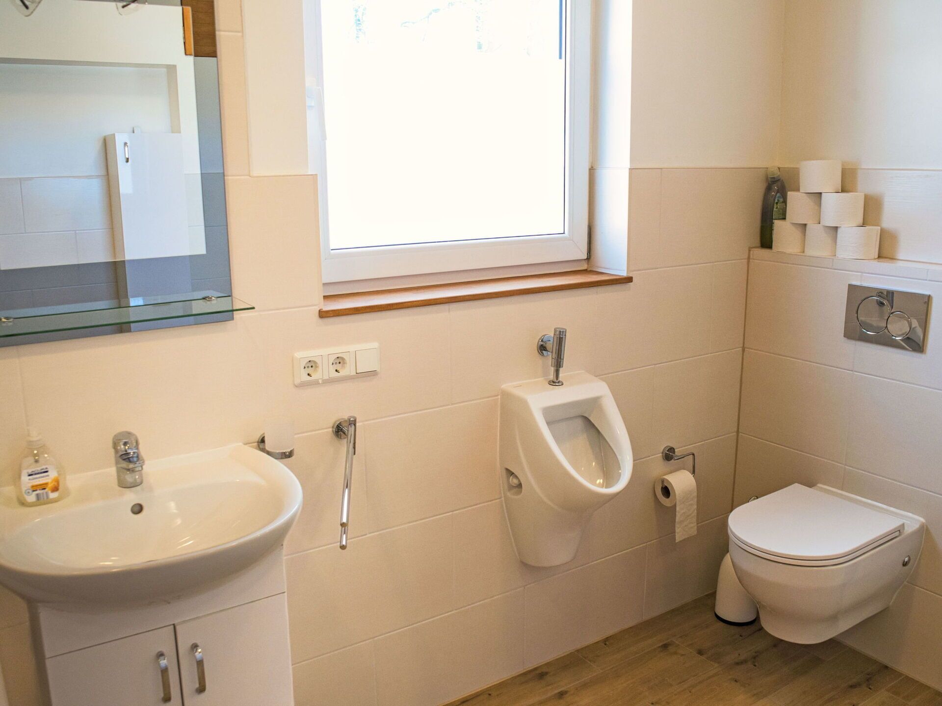 Une salle de bain moderne équipée d’un lavabo, d’un urinoir, de toilettes et d’une fenêtre laissant entrer la lumière naturelle.