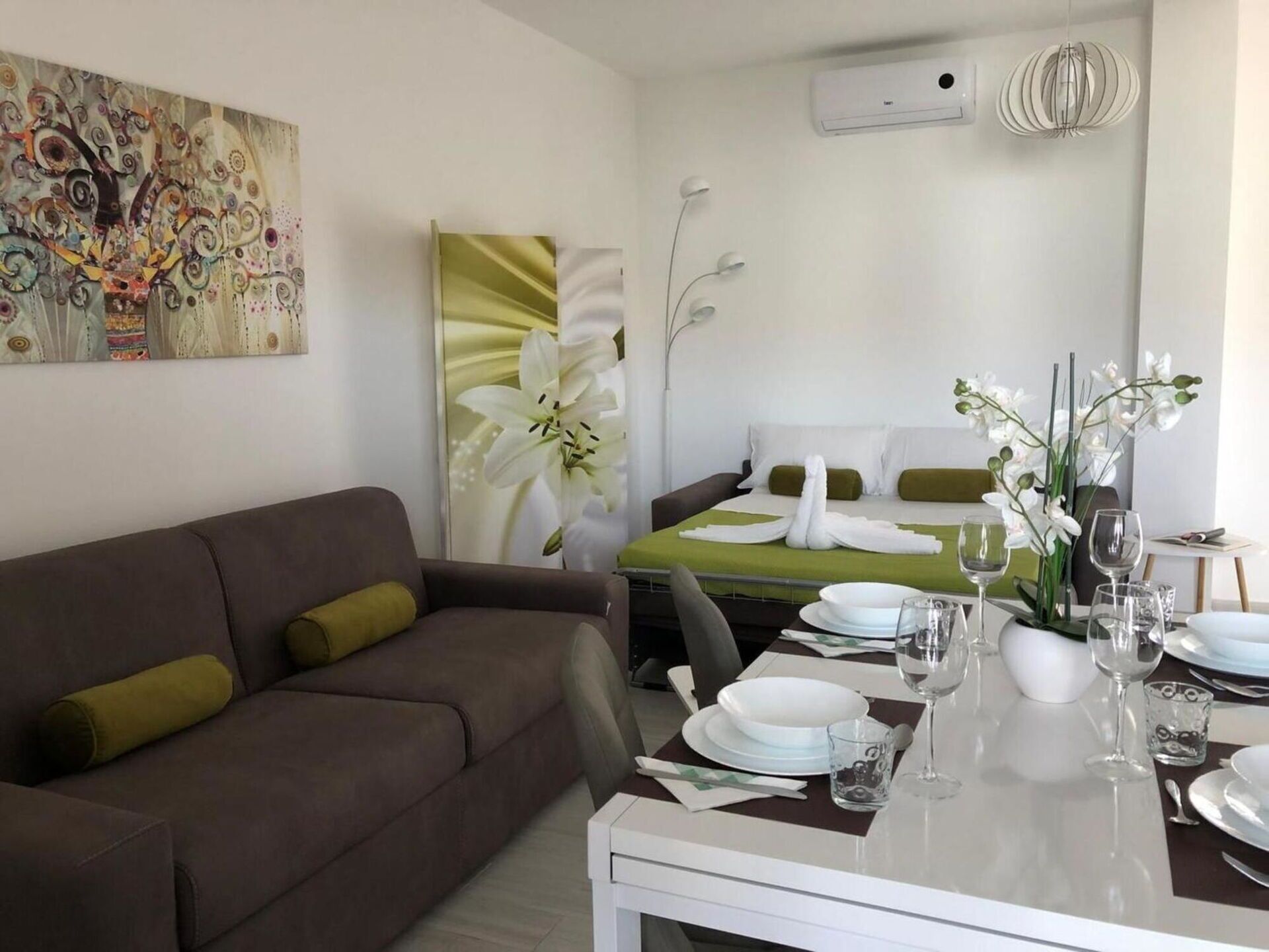 Moderni hotellihuone, jossa on ruskea sohva, siististi pedattu sänky, kahdelle katettu ruokapöytä ja suuri abstrakti maalaus seinällä.