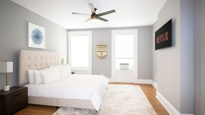 A bedroom with a tufted headboard bed, a ceiling fan, a framed artwork, a wall-mounted TV, and a large window with blinds. - 𖡡 Spring Suite in Hoboken - 1BR Apt - near NYC (Hoboken)