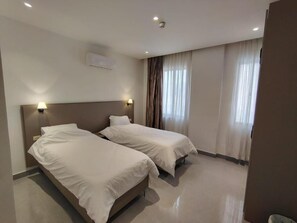 A hotel room with two beds, a ceiling light, an air conditioning unit, and a window with curtains.